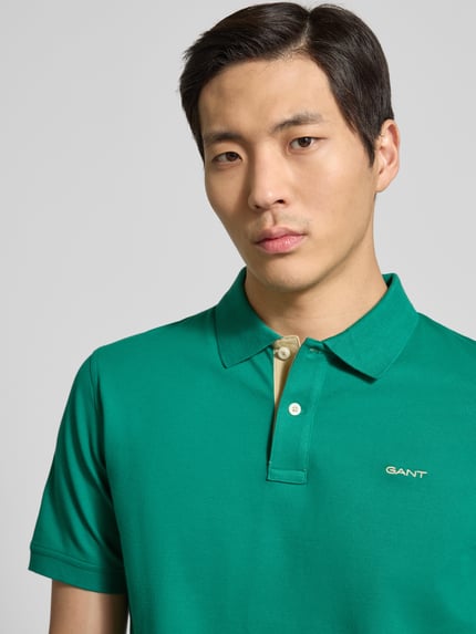 Męski model nosi Regular Fit Poloshirt mit Logo-Stitching od Gant, zielony kolor, elastyczna bawełna, krój regular fit, kołnierzyk polo, krótkie rękawy, zbliżenie na górną część ciała.