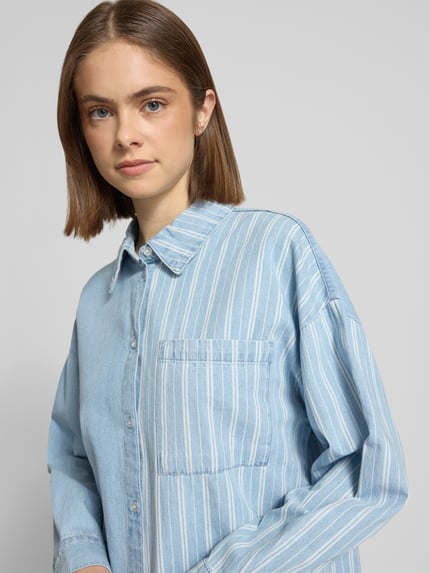 Modelka kobieca nosi Oversized Jeansbluse mit Brusttasche Modell 'KATRI' od Noisy May, jasnoniebieski kolor, bawełna, krój oversized, kieszeń na piersi, kołnierzyk koszulowy, listwa guzikowa, ramiona opuszczone, mankiety sportowe, zaokrąglony dół, długość pleców w rozmiarze S 72 cm, długość rękawa w rozmiarze S 50 cm, tekstura miękkiego denimu, widok z przodu, zbliżenie na górną część ciała.