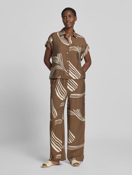 Frauenmodel trägt Regular Fit Hemdbluse aus Viskose-Mix Modell 'Zelyna' von someday, braun-beige mit abstraktem Muster, Kurzarm, Hemdblusenkragen, Knopfleiste, weiche Textur, Ganzkörperansicht von vorne.