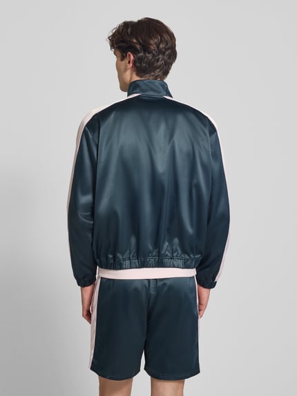 Männermodel trägt Schimmernde Trainingsjacke mit Label-Stitching von BLKVIS in dunklem Blau mit rosa Streifen, krój regular fit, efekt błyszczący, stójka, wyhaftowane logo, zamek błyskawiczny, długość tyłu w rozmiarze M: 61 cm, długość rękawów w rozmiarze M: 63 cm, Ganzkörperansicht Rückansicht