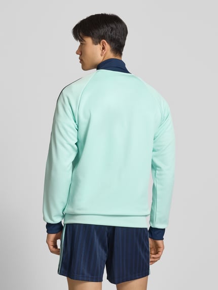 Mężczyznamodel nosi Regular Fit Trainingsjacke mit Baumwoll-Anteil und DFB-Logo von adidas Originals, miętowy kolor, długie rękawy, jednolita faktura, kontrastowe paski, boczne kieszenie, stehkraag, aplikacja DFB, krój regular fit, zamek błyskawiczny, widok całej sylwetki z tyłu.