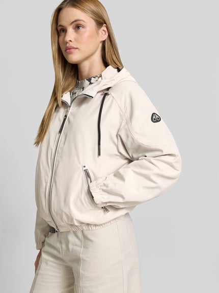 Frauenmodel trägt Jacke mit Zweiwege-Reißverschluss von Moose Knuckles in Beige mit Kapuze, Tunnelzug, Relaxed-Fit, Eingrifftaschen mit Reißverschluss, Rückenlänge 56 cm, Brustweite 56 cm, Ärmellänge 44 cm, Ganzkörperansicht von vorne.
