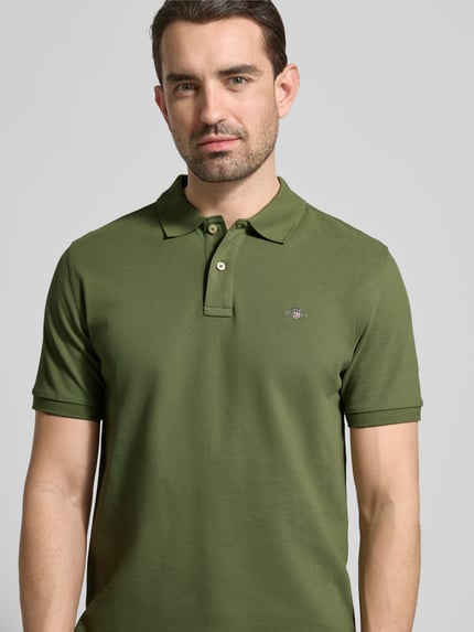Männermodel trägt Slim Fit Poloshirt mit Logo-Stitching von Gant aus Reine Baumwolle in Olivgrün, Nahaufnahme Oberkörper.