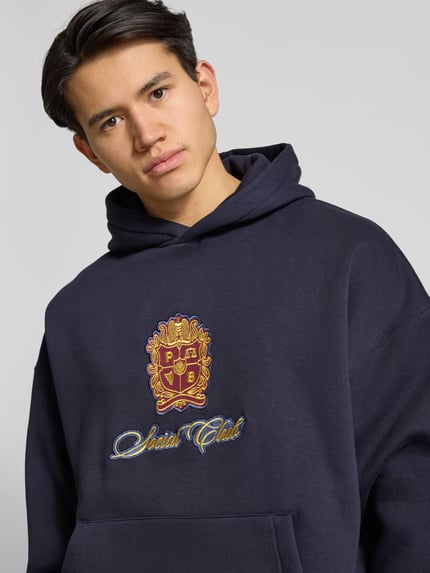 Männermodel trägt Hoodie mit Kapuze von PROHIBITED, dunkelblau, Stickerei vorn, Baumwoll-Mix, lockerer Schnitt, Nahaufnahme Oberkörper.
