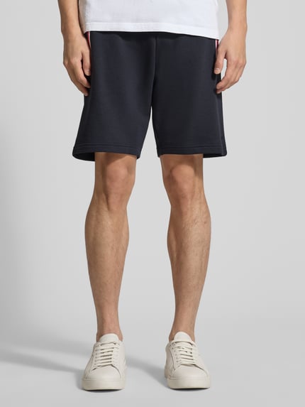 Mężczyzna nosi Shorts mit elastischem Bund von Christian Berg Men, bawełna z domieszką elastanu, krój regular fit, granatowe, kieszenie z kontrastowymi lamówkami, szerokość w rozmiarze M 43 cm, długość nogawki wewnętrznej 23 cm, widok całej sylwetki z przodu.