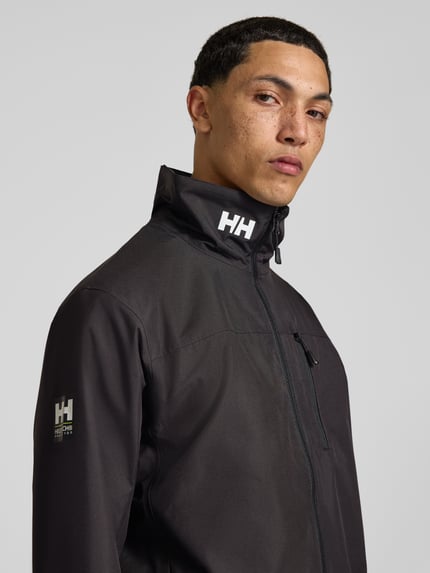 Männermodel trägt Regular Fit Jacke mit Stehkragen Modell 'Crew' von Helly Hansen in Schwarz, Logo-Print, Nahaufnahme Oberkörper.