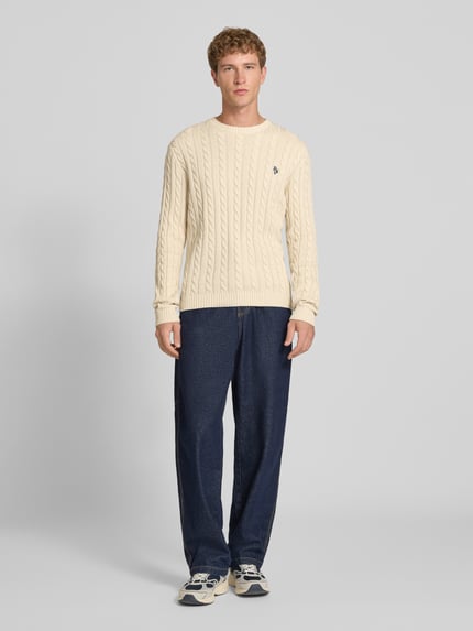 Mężczyzna nosi Slim Fit Strickpullover reine Baumwolle od REVIEW, kremowy sweter z dzianiny, krój slim fit, Ścieg warkoczowy, wyhaftowane logo, prążkowane ściągacze, długość tyłu w rozmiarze M: 73 cm, Długość rękawów w rozmiarze M: 70 cm, bawełna, Ganzkörperansicht von vorne.
