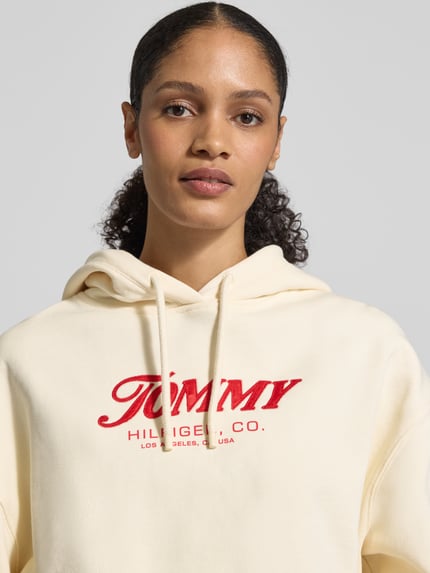 Vrouwenmodel draagt Regular Fit Hoodie aus reiner Baumwolle van Tommy Hilfiger in effen crème, labelprint, katoenen textuur, close-up bovenlichaam.