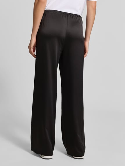 Männermodel trägt Wide Leg Stoffhose aus Satin von Juvia, schwarz, lockere Passform, elastischer Bund, Eingrifftaschen, Ganzkörperansicht Rückansicht