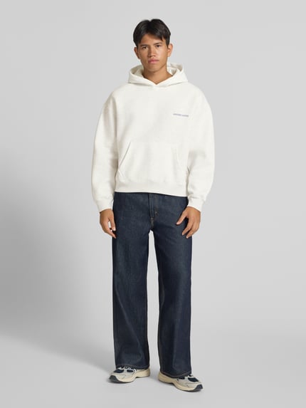 Männermodel trägt Relaxed Fit Hoodie aus Baumwoll-Mix Modell 'Sunday' von ANOTHER COTTON LAB, weiß, dunkle Jeans, Känguru-Tasche, elastische Abschlüsse, Ganzkörperansicht von vorne.