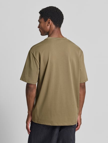 Męski model nosi Oversized T-Shirt aus reiner Baumwolle von Lindbergh, kolor oliwkowy, miękka faktura, długość tyłu w rozmiarze M: 70 cm, krój oversized, czysta bawełna, lekko opadające ramiona, półrękawkowy design, Ganzkörperansicht Rückansicht.