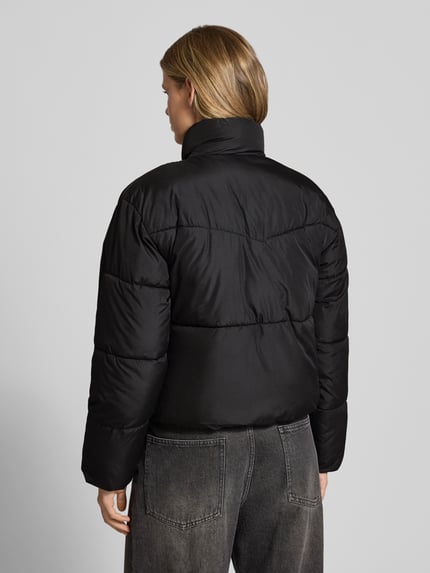 Frauenmodel trägt Steppjacke mit Stehkragen und Eingrifftaschen von JJXX in Schwarz, glatte Textur, Ganzkörperansicht Rückansicht. Regular Fit, Reißverschluss, Rückenlänge 58 cm, Schulterbreite 52 cm, Brustweite 58 cm, Ärmellänge 60 cm.