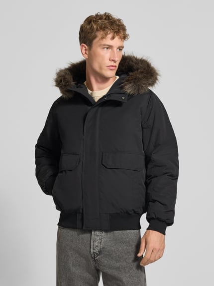 Männermodel trägt schwarze Bomberjacke mit Label-Patch Modell 'Everest' von Superdry, Kunstfellkapuze, Ganzkörperansicht von vorne. Regular Fit mit Reiß- und Druckknopfverschluss, Pattentaschen, Maße laut Kontext.