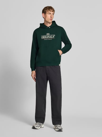 Männermodel trägt Hoodie mit Label-Print Modell 'FREDERIKSBERG' von JACK & JONES, dunkelgrün, lockerer Schnitt, Ganzkörperansicht von vorne. Baumwoll-Mix, Kapuze, Kängurutasche, gerippte Abschlüsse, Rückenlänge M 65 cm, Ärmellänge M 67 cm.