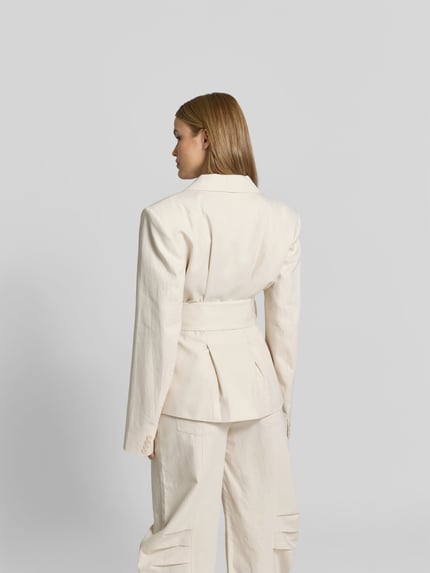 Männermodel trägt Blazer aus Leinen-Mix mit Taillen-Gürtel von ROTATE, kolor kremowy, krój slim fit, pasek w talii, długość tyłu 69 cm, szerokość w rozmiarze 36: 41 cm, długość rękawa 69 cm, szerokość talii 31 cm, textura gładka, widok całej sylwetki z tyłu