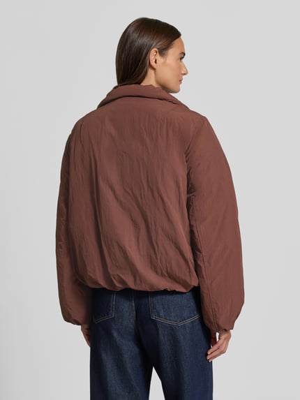 Frauenmodel trägt Jacke mit Stehkragen Modell 'TRUFA' von MANGO in Braun, strukturierte Oberfläche, gerader Schnitt, Ganzkörperansicht Rückansicht.