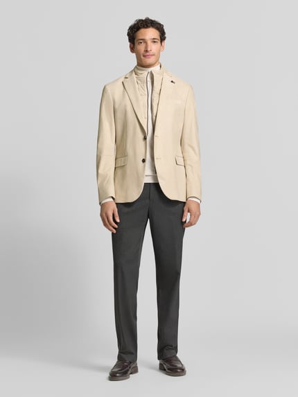 Męski model nosi Regular Fit Sakko aus elastischem Material-Mix mit Label-Detail von bugatti, beżowa elastyczna mieszanka materiałów, stójka, zamek błyskawiczny, marynarka na 2 guziki, kołnierz z połami, Brustasza, kieszenie z patką, długość tyłu w rozmiarze 50: 74 cm, szerokość ramion w rozmiarze 50: 43 cm, długość rękawów w rozmiarze 50: 66 cm, Ganzkörperansicht von vorne.