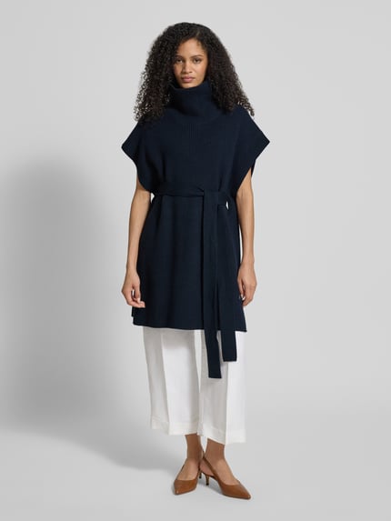 Frauenmodel trägt Regular Fit Poncho aus reiner Schurwolle Modell 'FAVANDA' von BOSS, Optik: unifarben, Strickoptik, Bindegürtel, Rollkragen, ohne Ärmel, Logo-Applikation, Materialverarbeitung: Strick, Ganzkörperansicht von vorne.