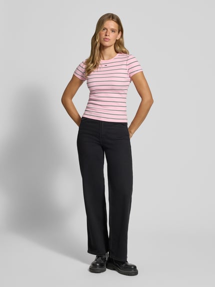 Frauenmodel trägt Slim Fit T-Shirt aus Baumwoll-Mix von Tommy Jeans in Rosa mit dunklen Streifen, schwarze Hose, Ganzkörperansicht von vorne. Damen T-Shirt von Tommy Jeans.