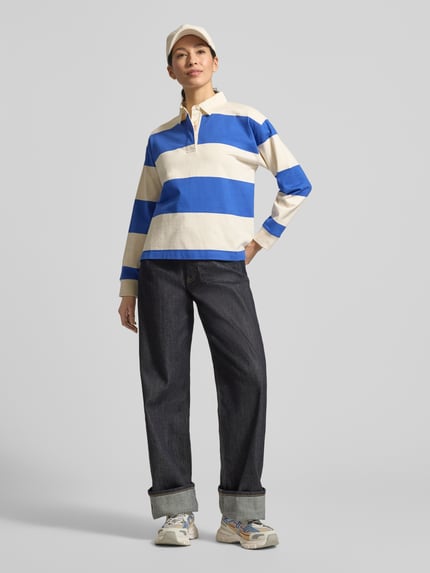 Frauenmodel trägt Regular Fit Langarm-Poloshirt mit verdeckter Knopfleiste von Gant in Blau-Weiß, reine Baumwolle, lockere Passform, Polokragen, sichtbare Seitenschlitze, kombiniert mit lässiger Hose, Ganzkörperansicht von vorne.