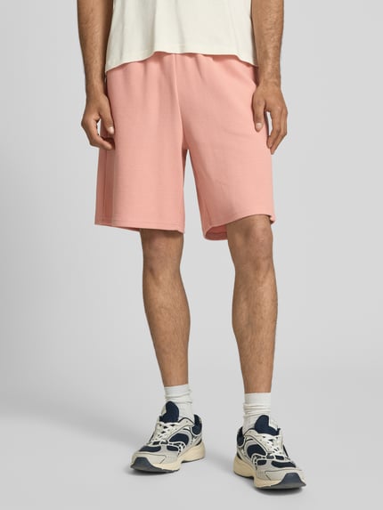 Männermodel trägt Regular Fit Shorts mit Strukturmuster von KARL KANI aus Baumwoll-Mix in Rosa mit Logo-Stitching, elastischer Bund, Ganzkörperansicht von vorne