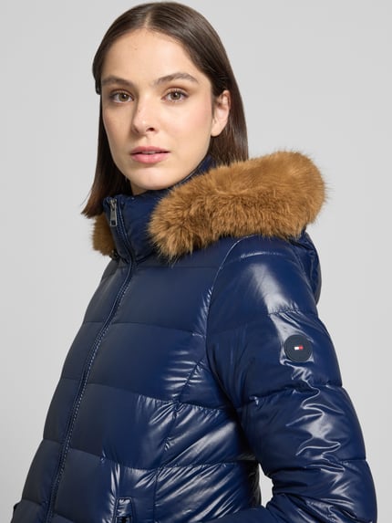 Frauenmodel trägt Puffer Coat mit abnehmbarer Fellkapuze von Tommy Hilfiger, glänzende Longline-Optik, Label-Detail, Nahaufnahme Oberkörper.