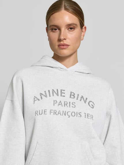 Frauenmodel trägt Oversized Hoodie aus reiner Baumwolle von Anine Bing in Hellgrau mit Label-Print, Känguru-Tasche, Nahaufnahme Oberkörper.