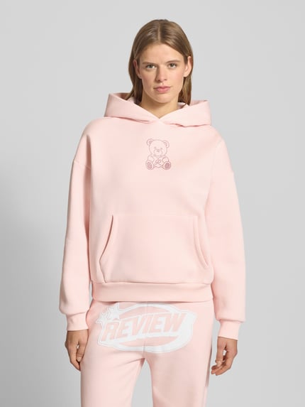 Frauenmodel trägt Hoodie mit Kapuze von Review in Rosa, Ziersteinbesatz, Känguru-Tasche, gerippte Abschlüsse, lockerer Schnitt, Ganzkörperansicht von vorne.