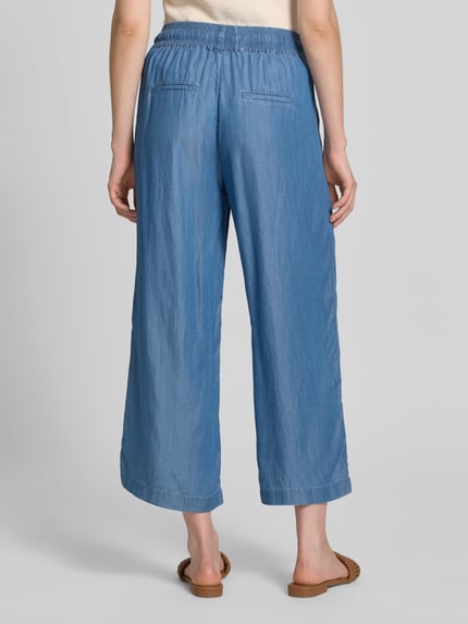 Frauenmodel trägt Wide Fit Stoffhose aus reinem Lyocell in Denim-Optik von Soyaconcept, helle Blautöne, verkürzte Passform, elastischer Bund mit Tunnelzug, Ganzkörperansicht Rückansicht.