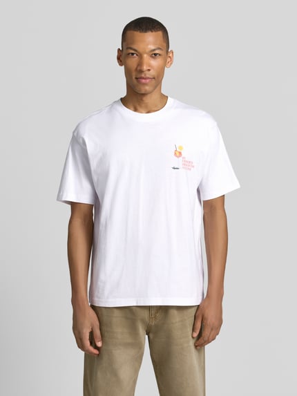 Męski model nosi T-Shirt mit rückseitigem Motiv-Print od MCNEAL, biała organiczna bawełna, krój regular fit, prosty fason, widok całej sylwetki z przodu.