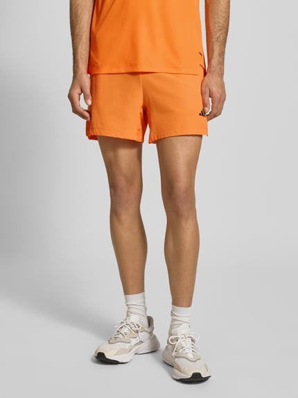 Männermodel trägt Regular Fit Shorts mit Logo-Print Modell 'Power' von adidas Training in Orange mit Strukturmuster, Eingrifftaschen, Nahaufnahme Oberschenkel vorne.