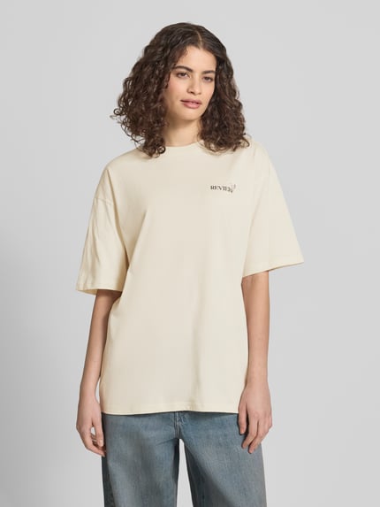Kobiece modelka nosi T-Shirt im Oversize-Fit mit Blumen-Backprint od REVIEW z czystej bawełny, krój oversized fit, okrągły dekolt, rękaw o dł. 1/2, kremowy, detal z logo, długość tyłu w rozmiarze S: 67 cm, widok na cały przód.