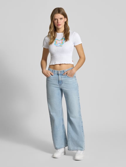 Frauenmodel trägt T-Shirt mit Label-Print von Levi's®, weiß, reine Baumwolle, körpernah, Rüschen, Smok-Details, Ganzkörperansicht von vorne.