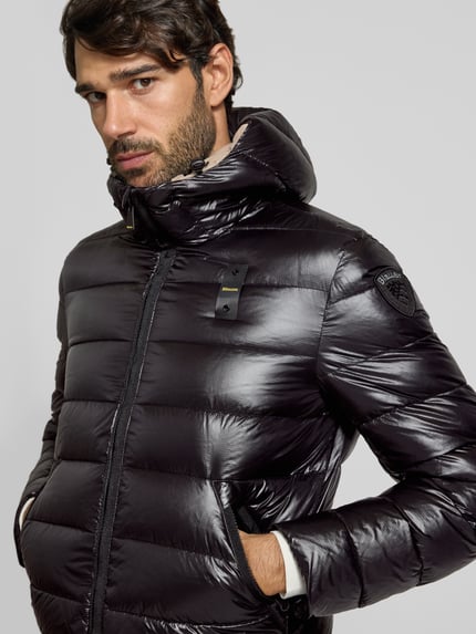 Männermodel trägt glänzende, schwarze Steppjacke mit Kapuze und Zweiwege-Reißverschluss Modell 'ADAMS' von Blauer USA, Nahaufnahme Oberkörper. Regular Fit, sichtbares Label-Detail, glatte Struktur.