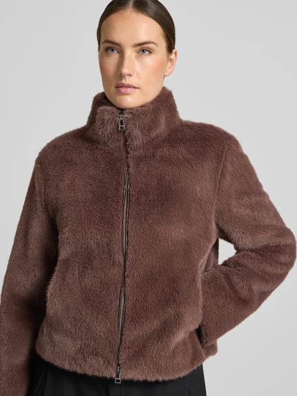 Frauenmodel trägt Jacke aus Kunstfell mit Zweiwege-Reißverschluss von Marc Cain in Braun, Stehkragen, flauschige Textur, Nahaufnahme Oberkörper, Regular Fit, KI-modifiziertes Bild.