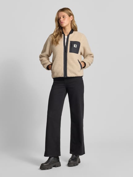 Frauenmodel trägt Fleecejacke mit Zweiwege-Reißverschluss Modell 'Janet' von Carhartt Work In Progress, Teddyfell, beige mit schwarzer Brusttasche, Regular Fit, sichtbare Eingrifftaschen und Stehkragen, Ganzkörperansicht von vorne.