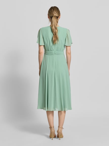 Frauenmodel trägt Knielanges Cocktailkleid mit Bindegürtel von Christian Berg Cocktail aus mintgrünem Chiffon, Taillierter Schnitt, Knielänge, Wickel-Optik, Rückansicht, Ganzkörperansicht Rückansicht. Rückenlänge bei Größe 36: 111 cm, Brustweite bei Größe 36: 38 cm, Ärmellänge bei Größe 36: 22 cm.