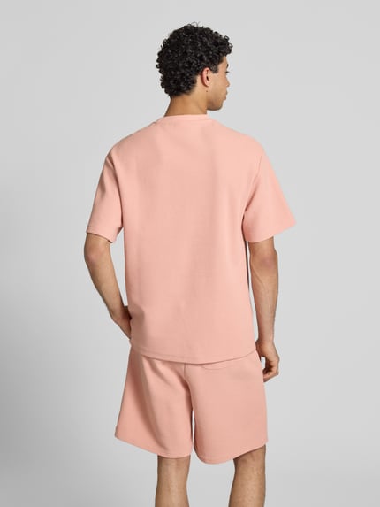 Männermodel trägt Oversized Fit T-Shirt mit Strukturmuster von KARL KANI, Baumwoll-Mix, hellrosafarbene Textur, Ganzkörperansicht Rückansicht.
