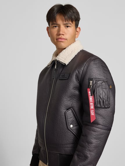 Männermodel trägt Bomberjacke mit Label-Patch und Kentkragen von Alpha Industries, schwarze Struktur, Teddyfellkragen, Nahaufnahme Oberkörper. Herren Bomberjacke, Regular Fit, Reißverschluss, Ärmeltasche mit Label-Detail, Eingrifftaschen mit Druckknopf, gerippte Abschlüsse.