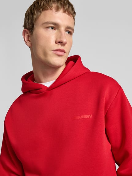 Männermodel trägt Regular Fit Hoodie mit Logo-Print von Review aus Baumwoll-Mix in kräftigem Rot, Nahaufnahme Oberkörper.