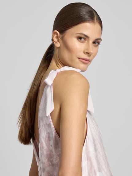 Frauenmodel trägt Relaxed Fit Kleid mit Allover-Paillettenbesatz von Jake*s Cocktail in Hellrosa mit Zierschleifen, Nahaufnahme Oberkörper.