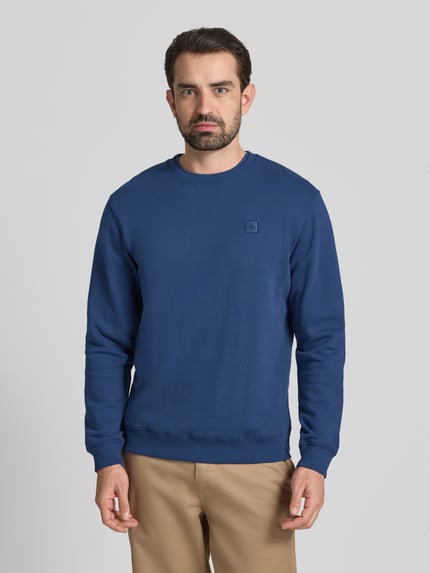 Männermodel trägt Sweatshirt mit Label-Detail von Scotch & Soda in Blau, Reine Baumwolle, Lockerer-Schnitt, Gerippte-Abschlüsse, Rundhalsausschnitt, Ganzkörperansicht von vorne.