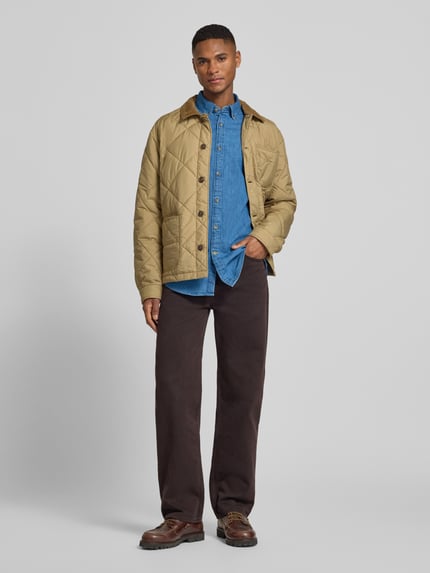 Männermodel trägt Slim Fit Jeanshemd aus reiner Baumwolle von Barbour in Denim-Optik, durchgehende Knopfleiste, Label-Stitching-Applikation, Langarm, Button-Down-Kragen, 1-Knopf-Manschette, leicht abgerundeter Rumpfabschluss, Farbe mittelblau, Ganzkörperansicht von vorne.