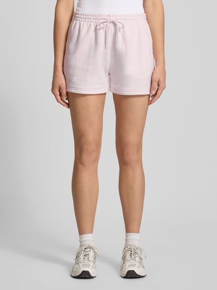 Frauenmodel trägt Straight Fit Shorts mit Tunnelzug Modell 'French' von New Balance aus Baumwoll-Mix in zartem Rosa, Ganzkörperansicht von vorne. Damen Shorts mit elastischem Bund, Bundweite bei Größe S: 40 cm, Innenbeinlänge: 8 cm.