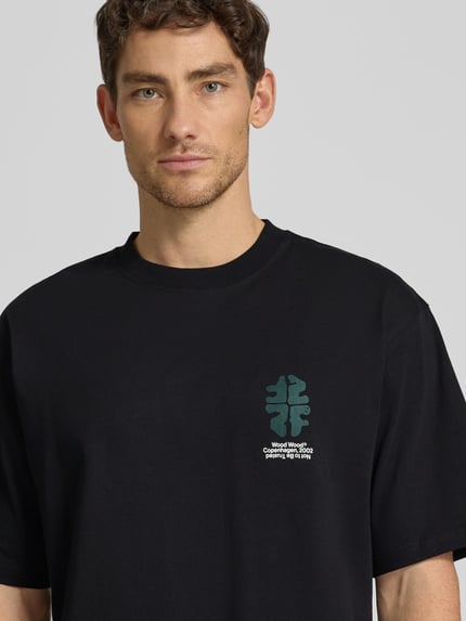 Männermodel trägt T-Shirt mit Rundhalsausschnitt Modell 'Gary' von Wood Wood aus reiner Baumwolle, Relaxed-Fit, Label-Print, Nahaufnahme Oberkörper.