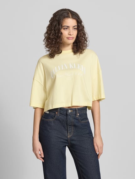 Vrouwenmodel draagt Cropped T-Shirt met logo-print van Calvin Klein Jeans in lichtgeel, puur katoen, korte fit, ronde hals, ruglengte maat S 42 cm, logoprint op borst, ganzekörperaanzicht van voren.
