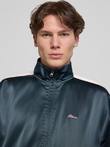 Męski model nosi Schimmernde Trainingsjacke mit Label-Stitching od BLKVIS, krój regular fit, efekt błyszczący, stójka, zamek błyskawiczny, długość tyłu w rozmiarze M: 61 cm, długość rękawów w rozmiarze M: 63 cm, kolor ciemny z jasnymi detalami, wyhaftowane logo, Nahaufnahme górnej części ciała.