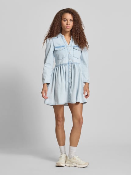 Frauenmodel trägt Mini-Jeanskleid aus reiner Baumwolle Modell 'CYRUS' von Only in heller Denim-Optik, Ausgestellt, Mini-Länge, V-Ausschnitt, Rückenpasse, Knopfleiste, Langarm mit schmaler Manschette, Ganzkörperansicht von vorne.