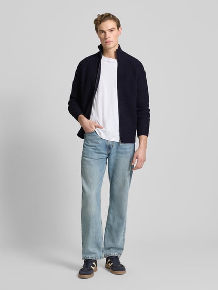 Männermodel trägt Regular Fit Strickjacke aus reiner Wolle von Forét in Dunkelblau, gerippter Stehkragen, Reißverschluss, Ganzkörperansicht von vorne.