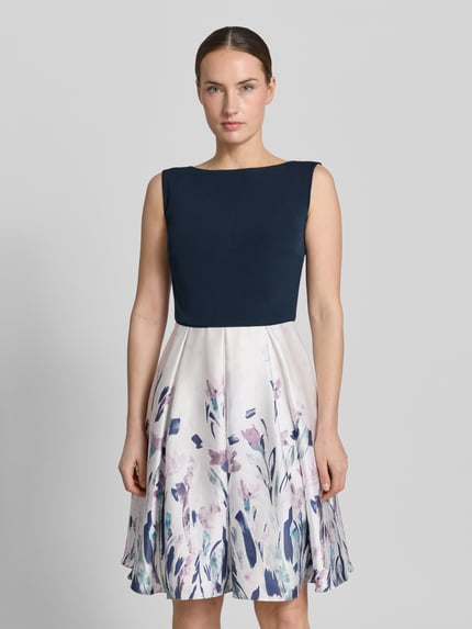 Frauenmodel trägt knielanges Cocktailkleid mit U-Boot-Ausschnitt von Christian Berg Cocktail, Satin, Taillierter Schnitt, Knielänge, U-Boot-Ausschnitt, Rückseitiger V-Ausschnitt, Rückseitiger Reißverschluss, Rückenlänge bei Größe 36: 75 cm, Brustweite bei Größe 36: 40 cm, dunkles Oberteil, heller Rock mit abstraktem Muster, Ganzkörperansicht von vorne.